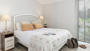 A bedroom with a bed, bedside lamps, a nightstand, a window with blinds, and a view of greenery outside. - 2 bedroom accommodation in Honiton (Honiton)