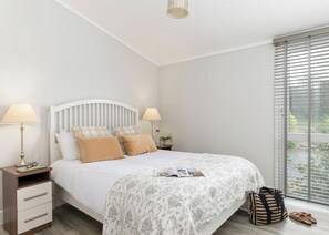 A bedroom with a bed, bedside lamps, a nightstand, a window with blinds, and a view of greenery outside.