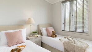 A bedroom with two beds, a nightstand, a lamp, a teddy bear, and a window with blinds. - 2 bedroom accommodation in Honiton (Honiton)