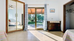 Una habitación con puerta corrediza de vidrio que da a un jardín, una cama con colcha a rayas, un aparador de madera y un ventilador de techo.