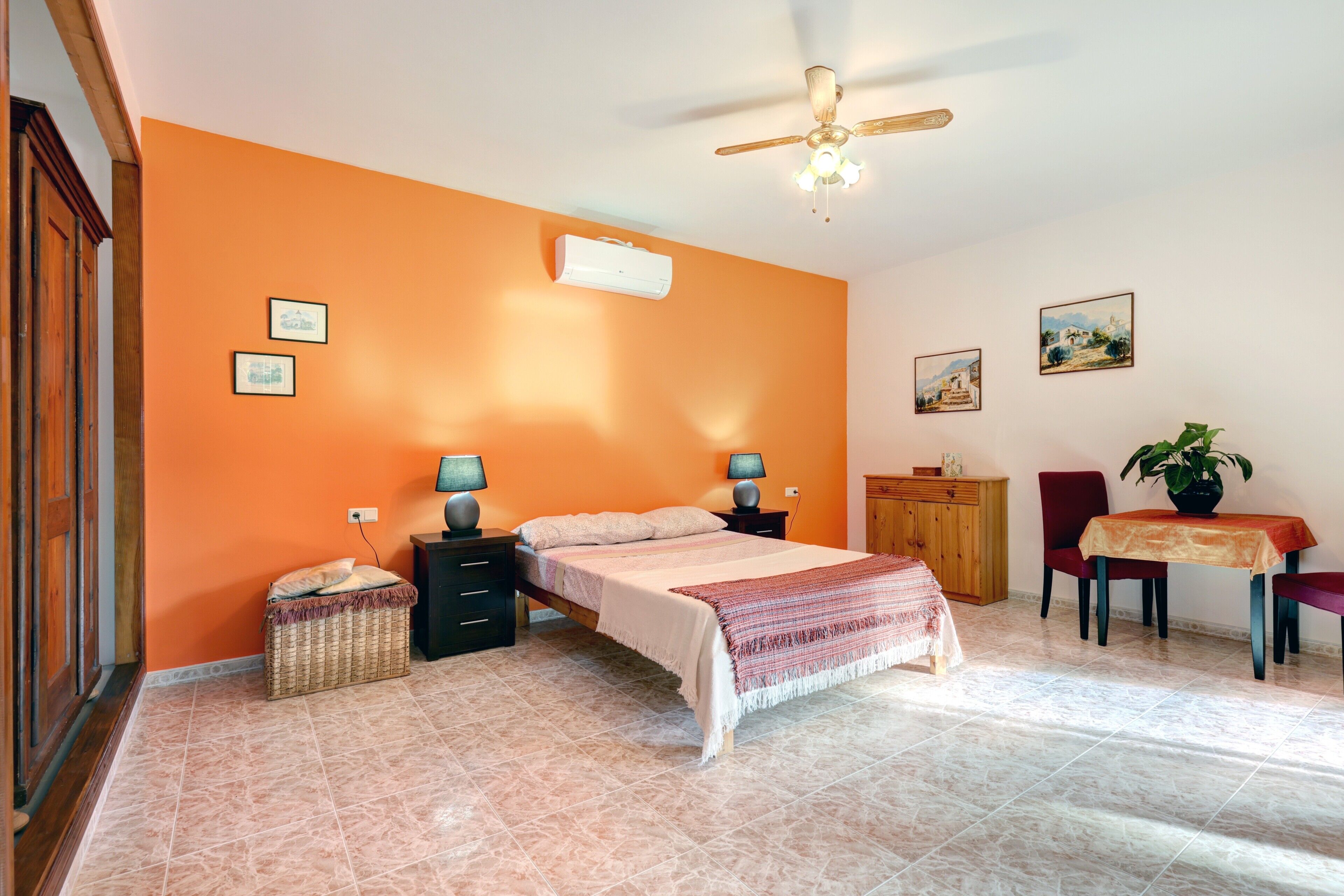 A bedroom with a bed, bedside tables, a ceiling fan, an air conditioner, a wooden cabinet, a dining table with chairs, and a potted plant.