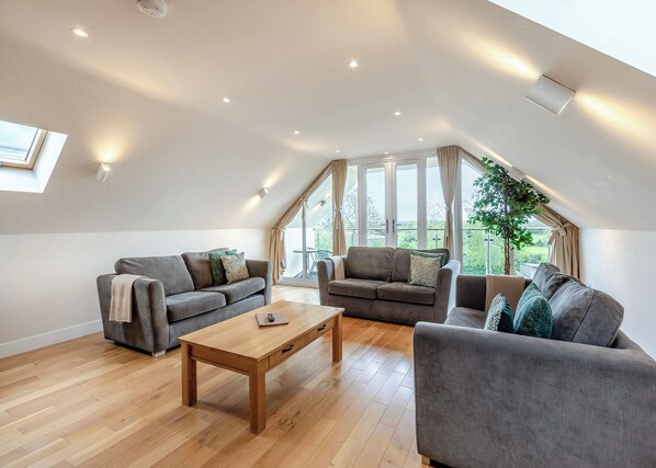 A spacious loft with a high ceiling, two grey sofas, a wooden coffee table, and large windows offering a view of greenery.