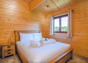 A wooden-paneled room with a bed, a nightstand, a window with curtains, and a ceiling light. - Luxurious solid log lodge ideal for couples. (Wenhaston, Halesworth)