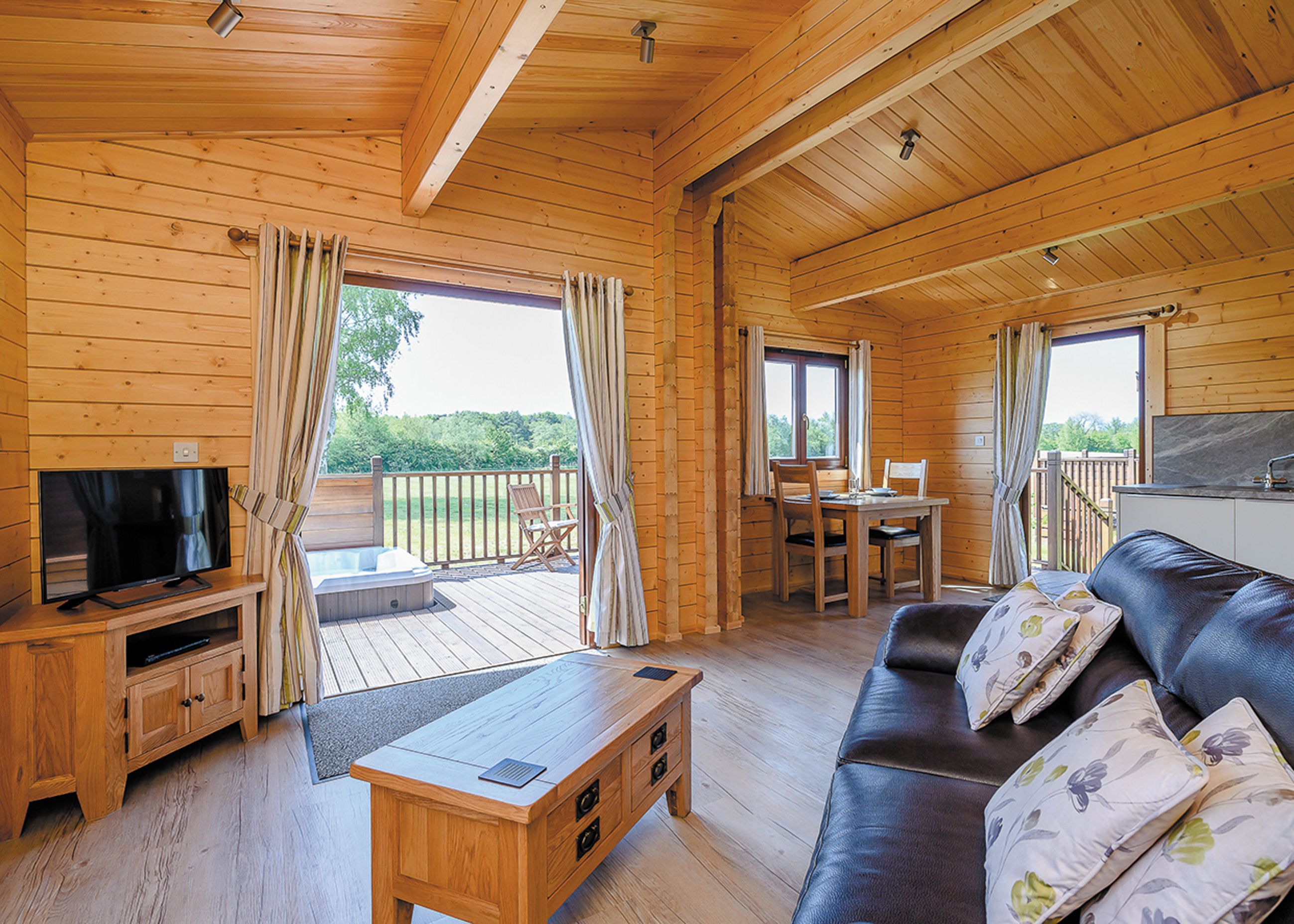 A wooden cabin interior with a TV, sofa, dining table, and hot tub.
