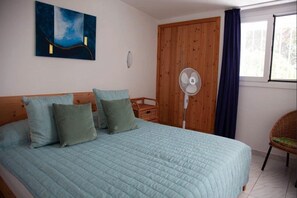 Une chambre à coucher avec un lit, une armoire en bois, un ventilateur sur pied, une chaise en osier et une fenêtre avec des rideaux.