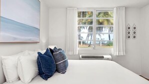 Ein Schlafzimmer mit einem großen Bett, weißen Vorhängen, einem Fenster mit Blick auf Palmen und einem an der Wand befestigten Bilderrahmen.