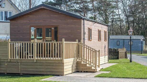 A small wooden cabin with a wooden deck, a staircase, and a fenced area.