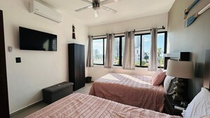 A hotel room with two beds, a ceiling fan, an air conditioner, a TV, a window with curtains, and a view of buildings outside.