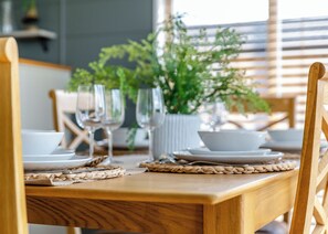 Una mesa de comedor de madera con platos, cuencos y vasos blancos, un protector de mesa tejido y una planta en maceta al fondo.