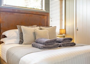 A neatly made bed with pillows and folded towels, a wooden headboard, and a window with blinds.