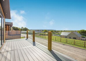 A wooden deck with a view of a rural landscape, including a stone building and a fenced area.
