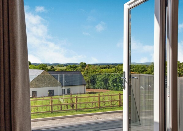 A balcony with a view of a rural landscape, a wooden fence, and a building with a sloped roof.