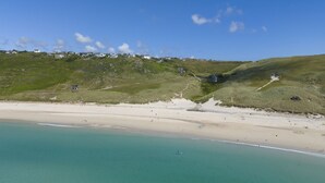 A coastal landscape with a sandy beach, clear turquoise waters, and a grassy hill with buildings.