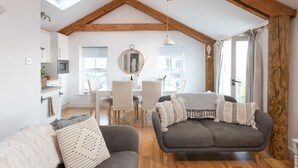 A spacious loft with exposed wooden beams, a cozy seating area with a sofa and armchairs, a dining table with chairs, and a kitchen area with a microwave.