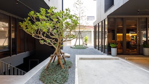 Um edifício moderno com um pátio, apresentando uma árvore, caminhos de cascalho e plantas em vasos.
