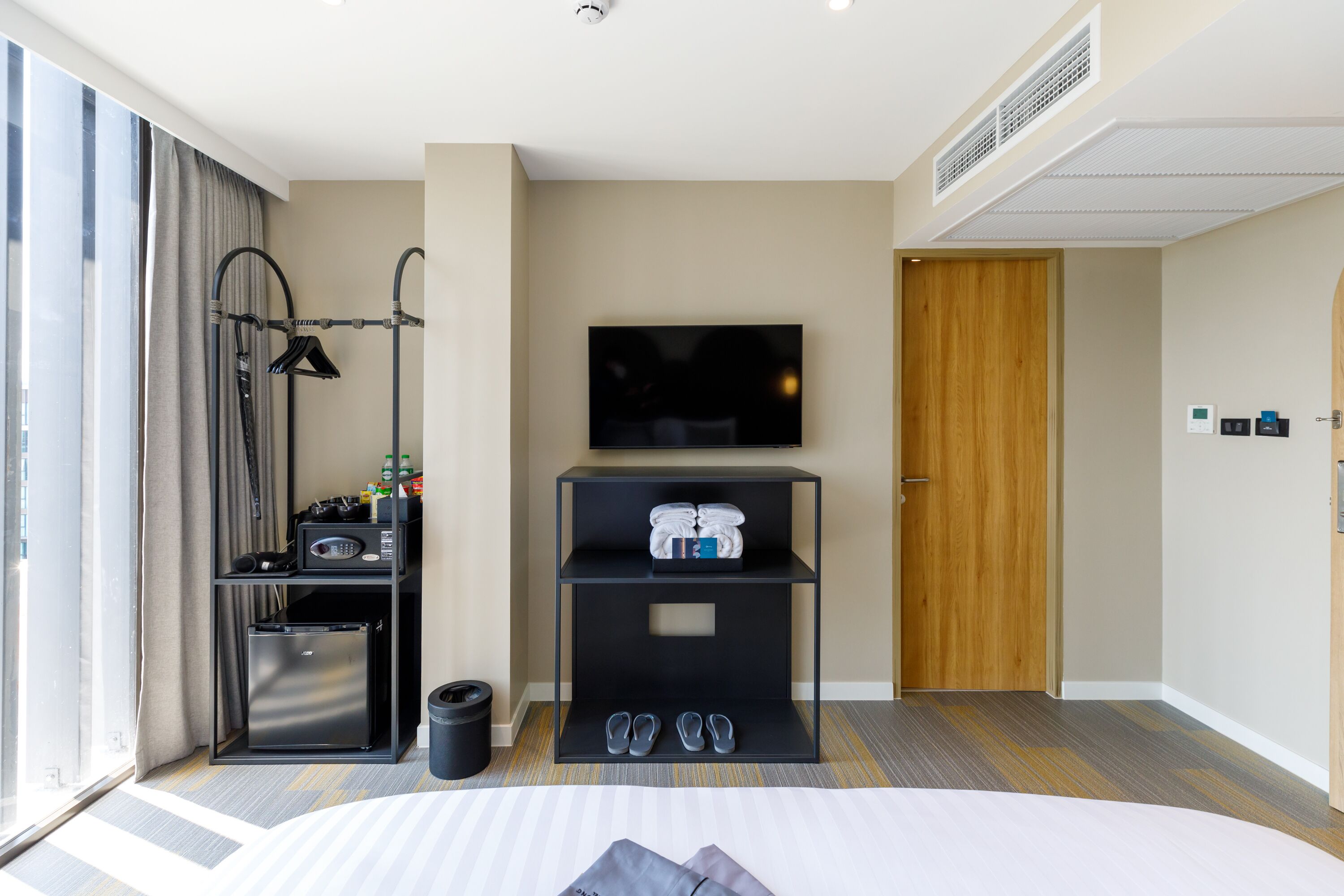A hotel room with a bed, a TV, a minibar, a shoe rack, and a door.