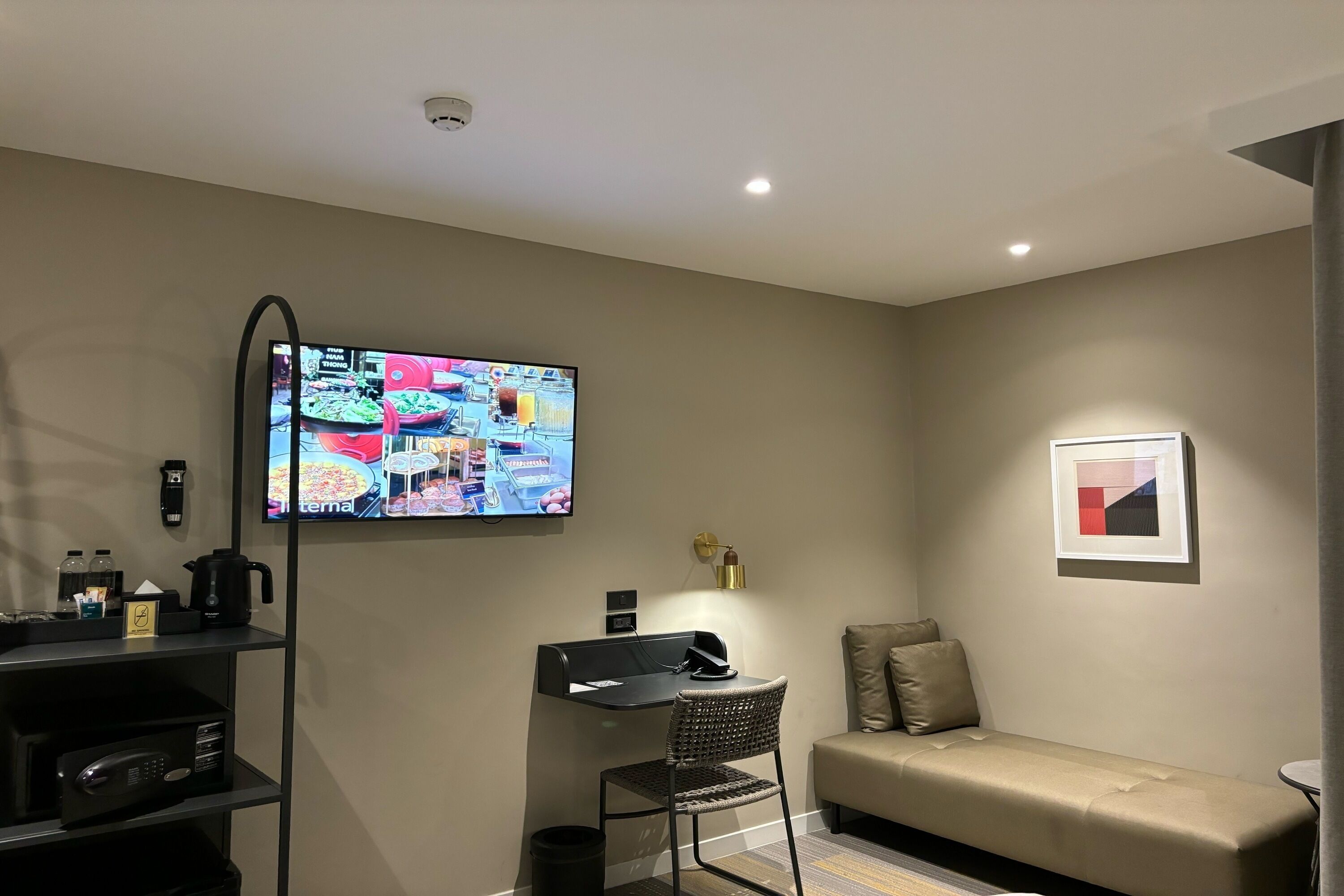 Ein modernes Zimmer mit einem wandmontierten Flachbildfernseher, einem schwarzen Schreibtisch mit Stuhl, einem beigen Sofa und einem kleinen Tisch mit Lampe.