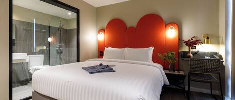 A hotel room with a large bed, a glass shower enclosure, a bedside table with a lamp, and a chair.