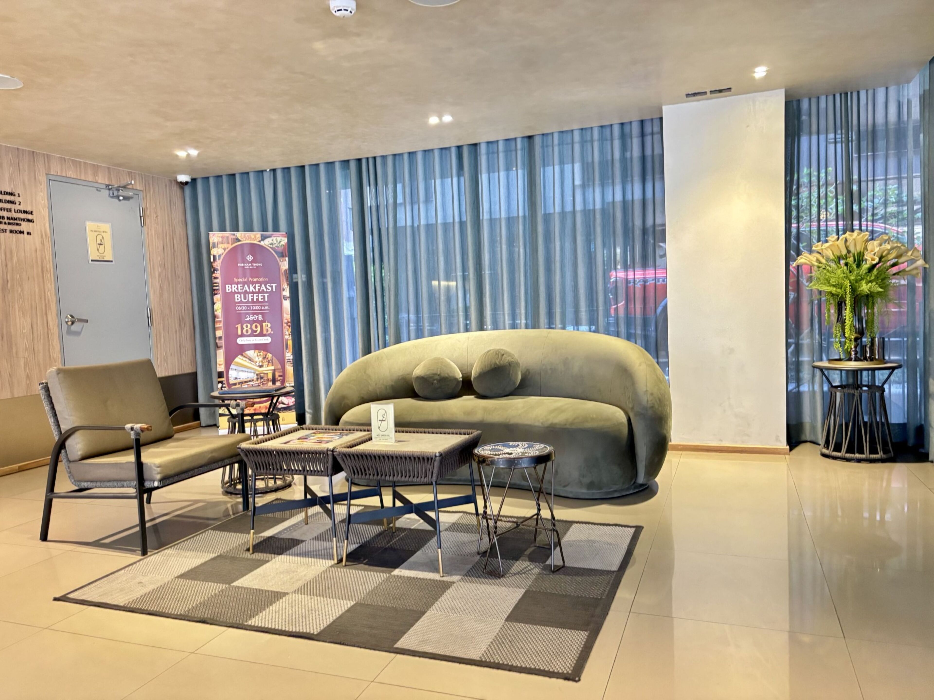 A modern hotel lobby with a plush sofa, a coffee table, and a chair. A large window with curtains is visible, and a signboard is present.