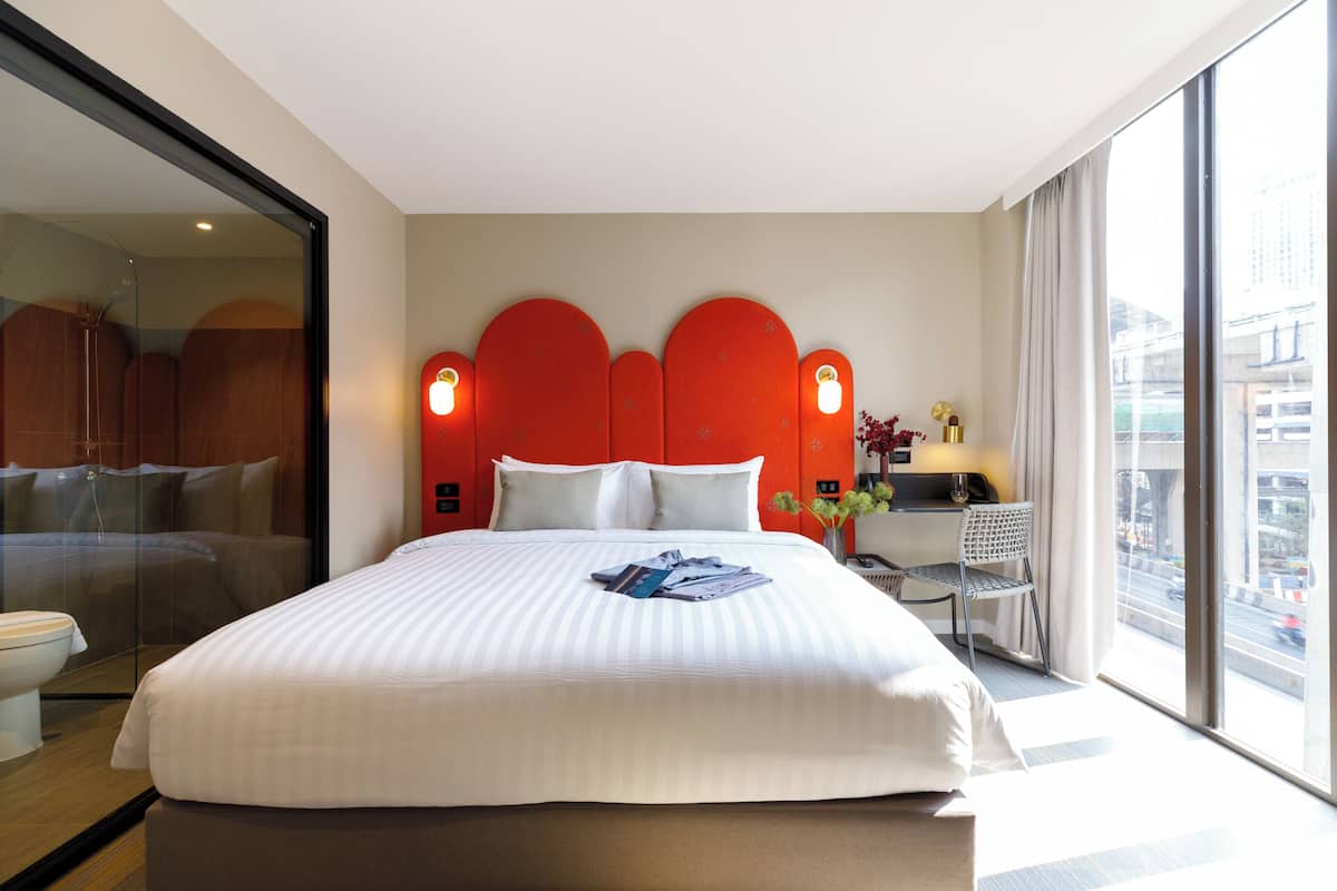 Een hotelkamer met een groot bed, een bureau met stoel en uitzicht op de stad door grote ramen.