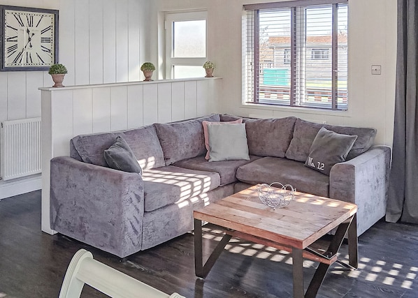 A living room with a sectional sofa, a wooden coffee table, a clock on the wall, and a window with blinds.