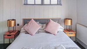 A neatly made bed with pillows, two bedside tables with lamps, a radiator, and a window with blinds. - 3 bedroom accommodation in Newbury (Newbury)