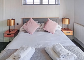 Una cama bien hecha con almohadas, dos mesitas de noche con lámparas, un radiador y una ventana con persianas.