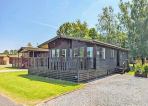 A wooden mobile home with a fenced deck, a gravel driveway, and a grassy lawn.