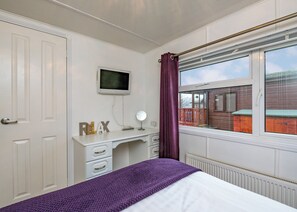 Quarto com cama, escrivaninha com computador e luminária, televisão na parede e janela com vista para uma casa.
