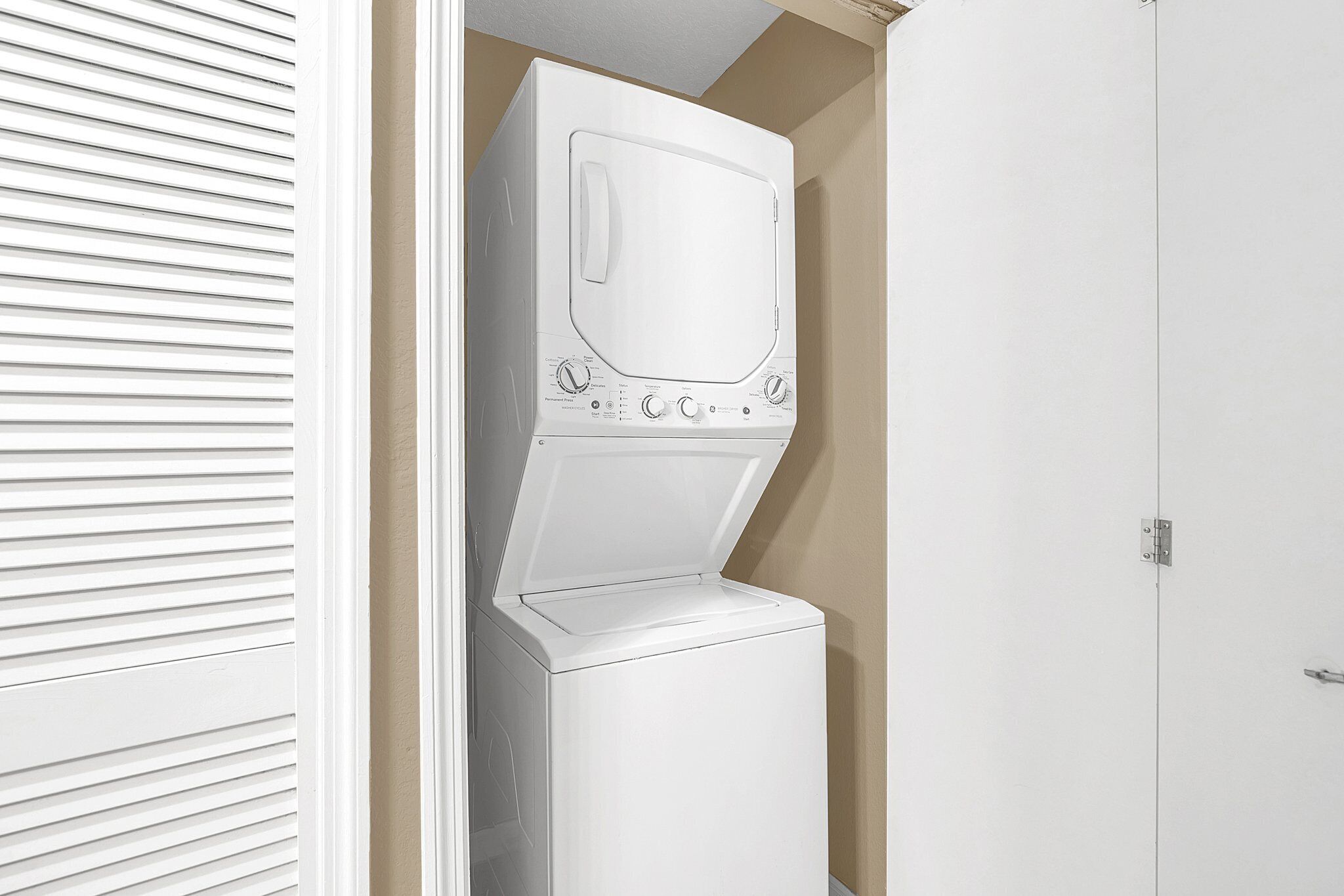 Una lavadora y secadora apilables, de color blanco y tamaño compacto, en un pequeño cuarto de lavado con pared beige y puerta blanca.