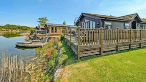 A lakeside wooden cabin with a dock, surrounded by a wooden fence, and a grassy area.