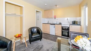 Een compacte leefruimte met een kitchenette, een glazen eettafel, een leren fauteuil en een kleine koelkast.
