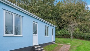 A blue building with a white door and windows, a small set of steps leading to the entrance, and a grassy yard with a tree.