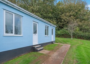 A blue building with a white door and windows, a small set of steps leading to the entrance, and a grassy yard with a tree.
