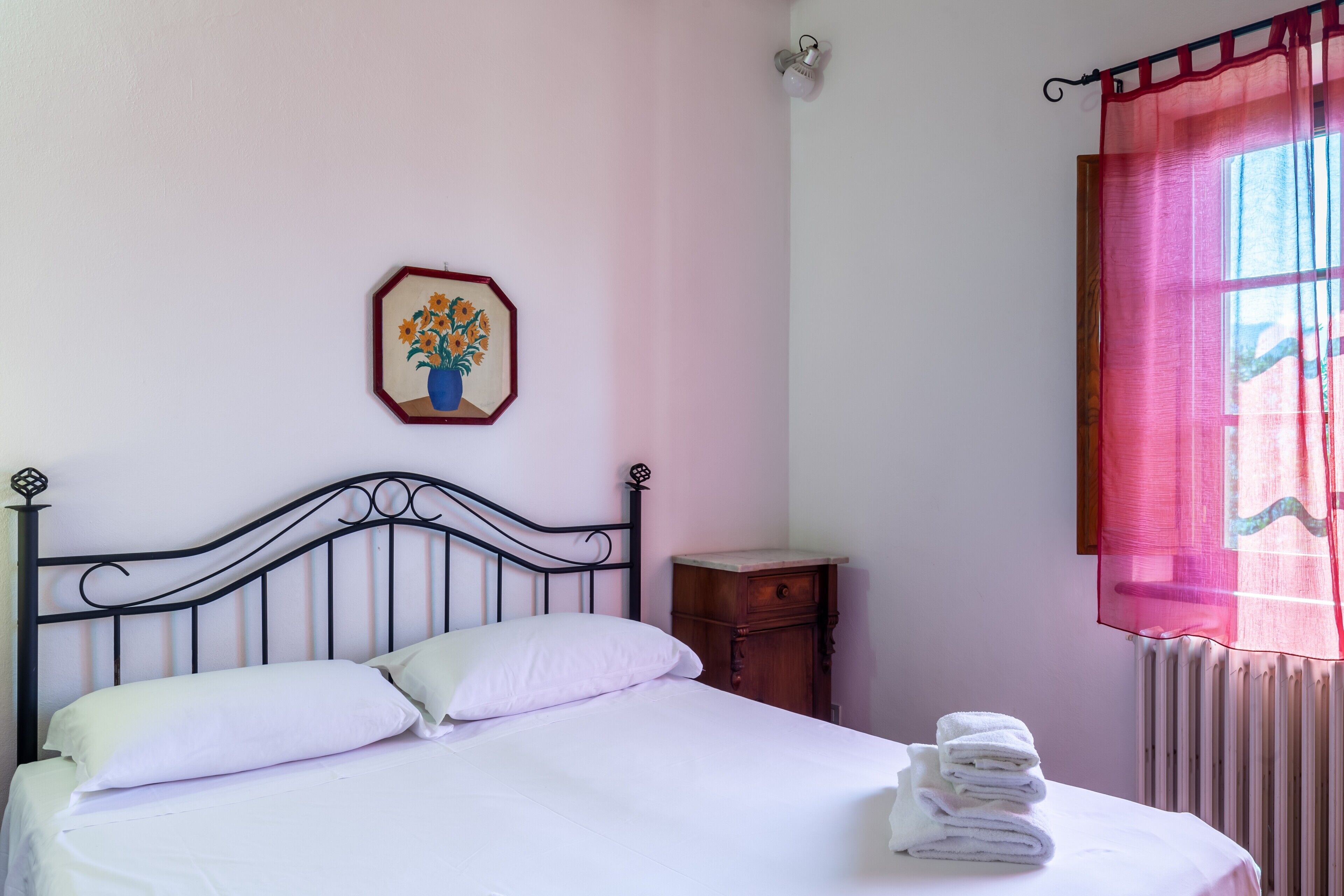 Un letto con lenzuola bianche, un comodino in legno, un quadro incorniciato e una finestra con tenda rosa.