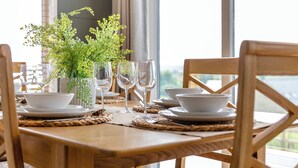 Une table de salle à manger en bois, dressée avec de l’assiette blanche, des bols et des verres à vin, agrémentée d’une plante verte dans un vase blanc.