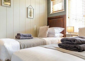 Une chambre d’hôtel avec deux lits, une tête de lit en bois, une table de chevet avec une lampe et une fenêtre avec des stores.