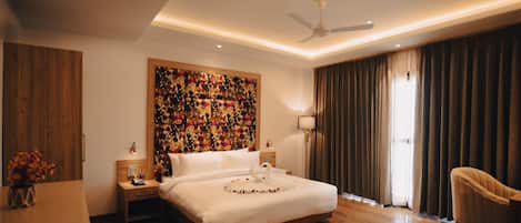 A modern hotel room with a large bed, wooden flooring, a ceiling fan, and a large window with curtains.