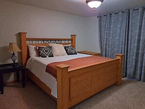 A wooden bed with a headboard, a bedside table with a lamp, and a window with curtains. - Big Pine Bluff (Perham)