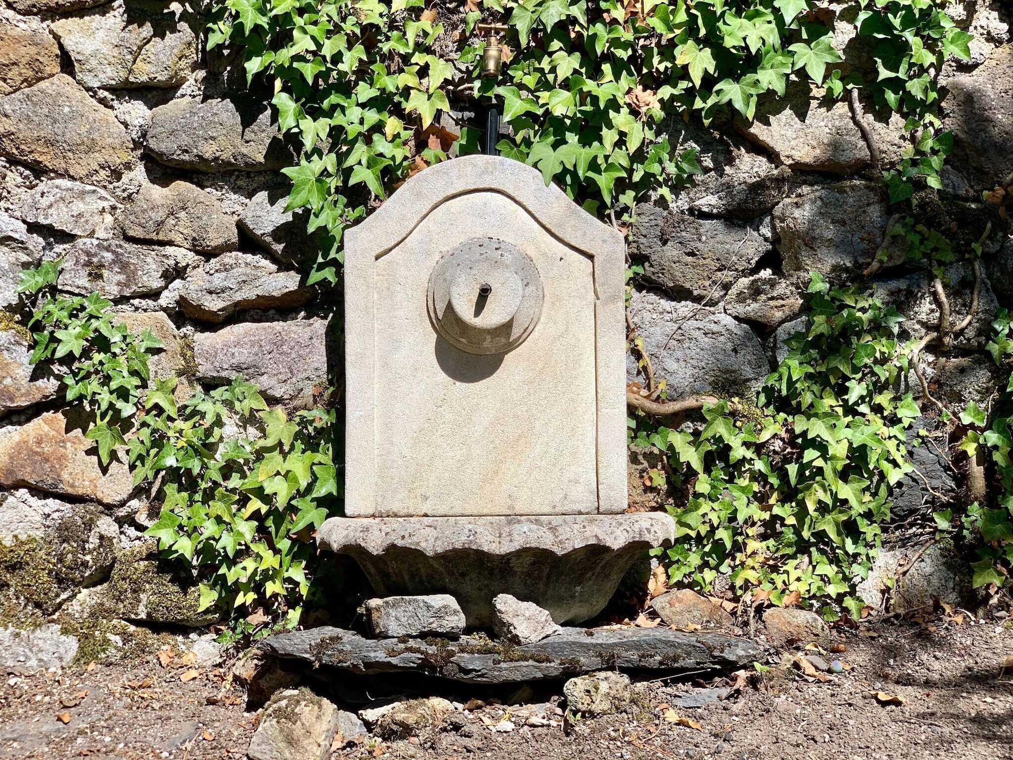 Un monument de pierre surmonté d’un élément circulaire, posé sur un socle en pierre, entouré d’un mur de pierre et de verdure.