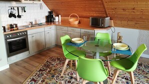 A compact kitchen with modern appliances, a dining area with a glass table and green chairs, and a patterned rug. - Tranquil Waterside Hideaway (Usedom)