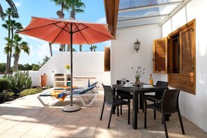 Un patio con una mesa, sillas, un paraguas rojo y una pared blanca con contraventanas de madera.