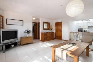 A modern living room with a flat-screen TV, wooden furniture, a dining table, and a kitchen area with cabinets and a large window.