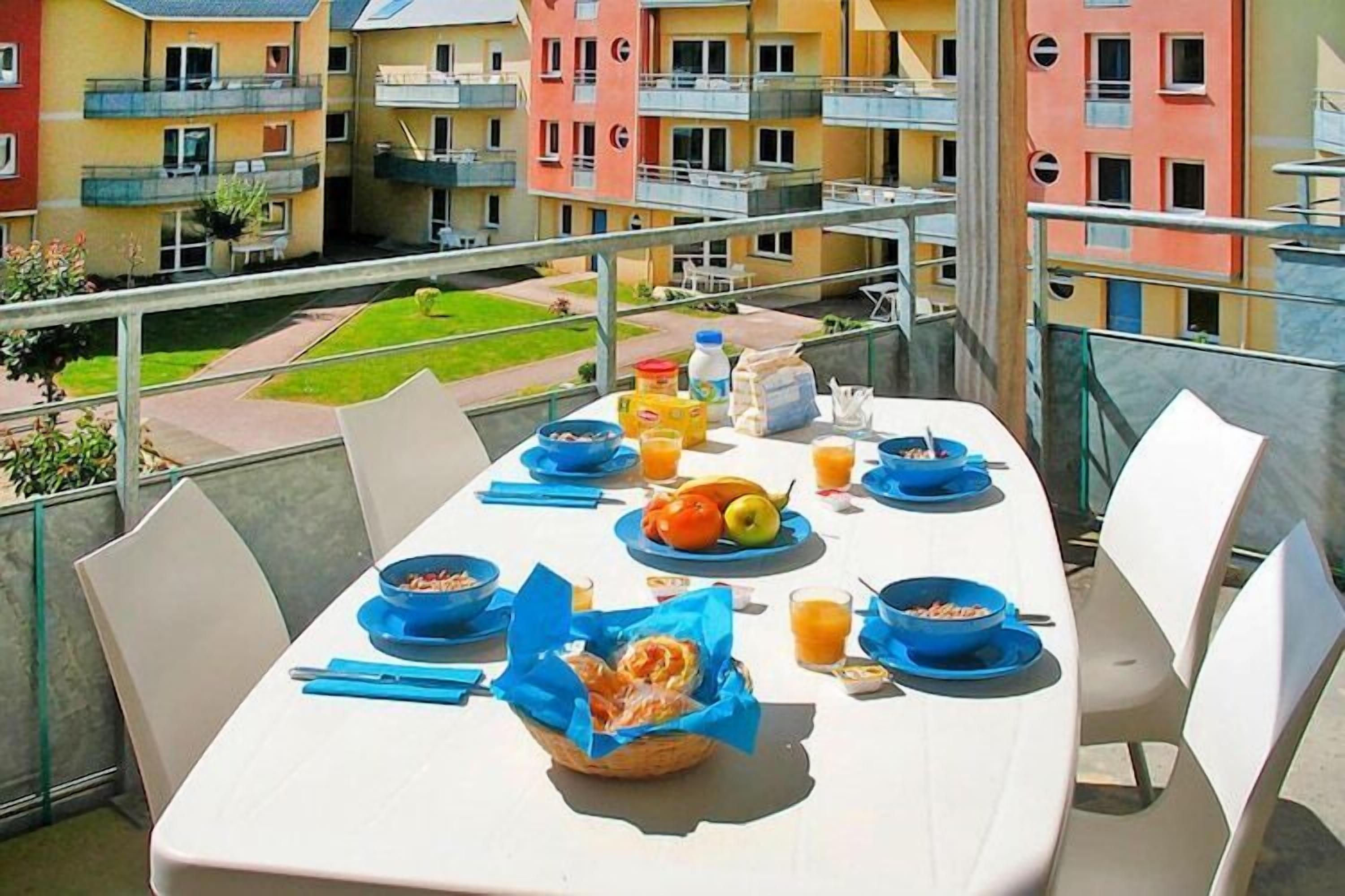 Une table de balcon dressée avec des éléments pour le petit-déjeuner, entourée d’immeubles d’habitation.
