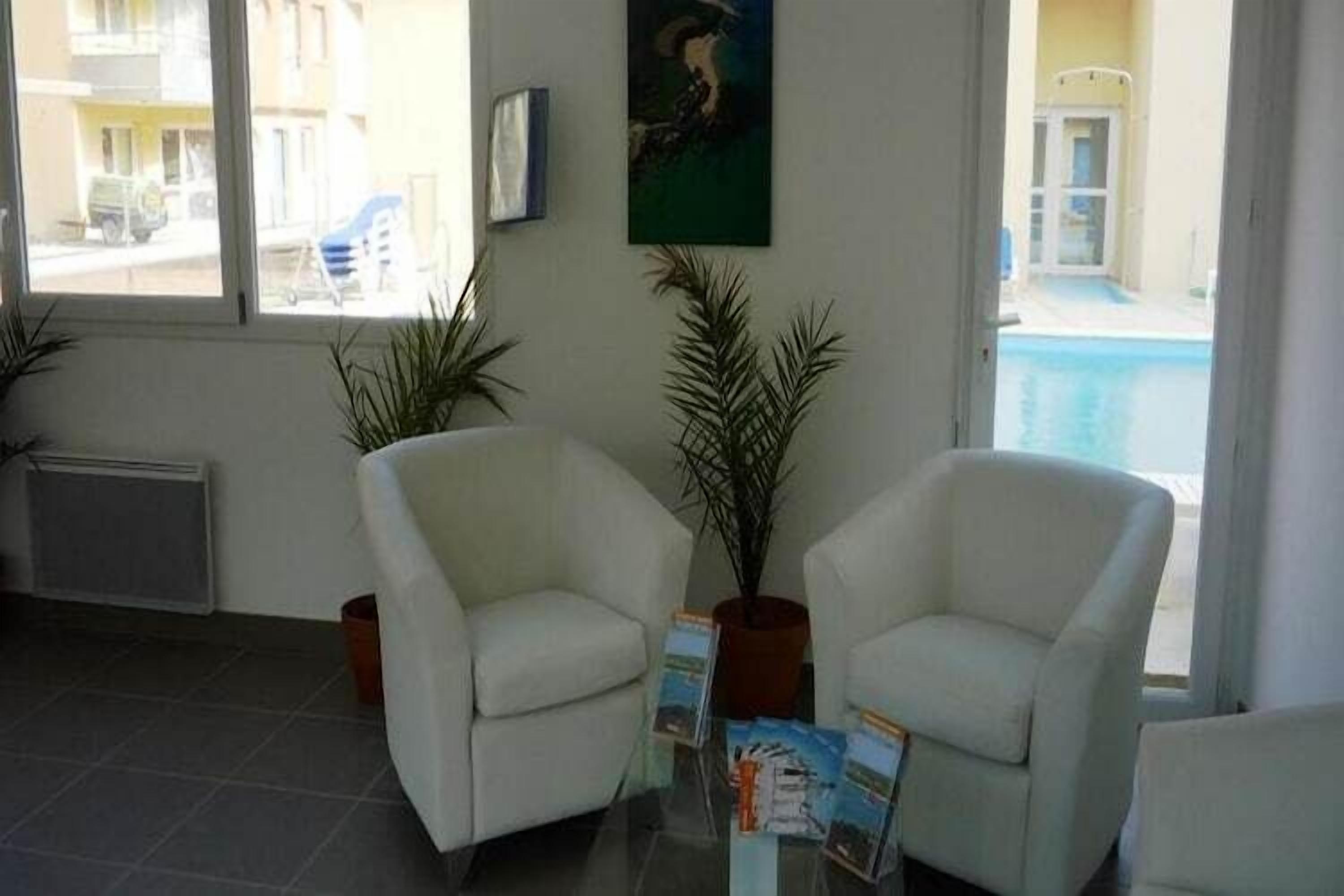A room with two white armchairs, a glass table with brochures, a potted plant, a painting on the wall, and a view of a swimming pool outside.