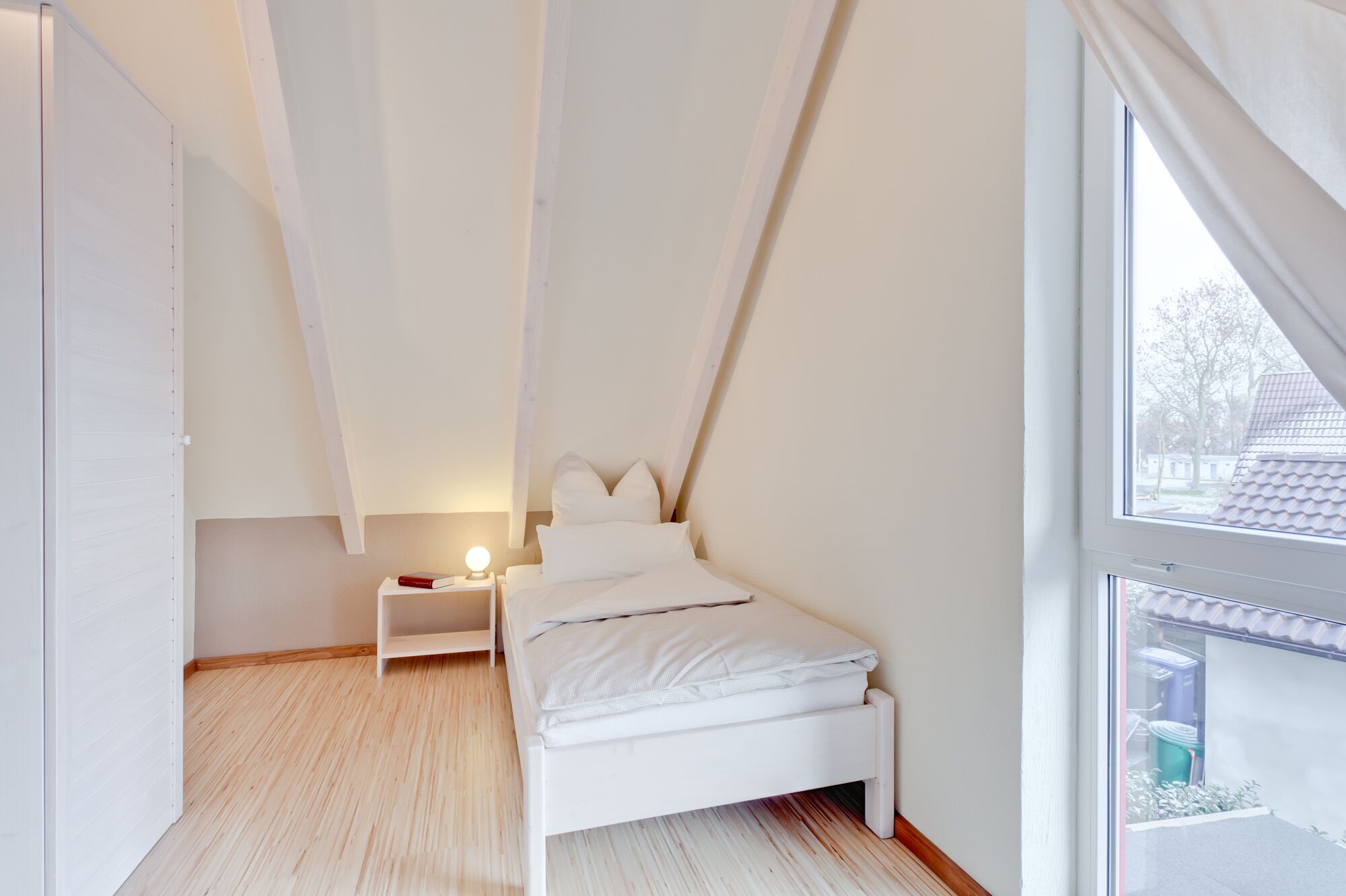 A bedroom with a sloped ceiling, a bed, a bedside table with a lamp, and a window with a view of the outside.