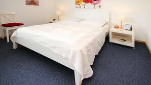 A neatly made bed with white bedding, a red chair with a red cushion, a nightstand with a lamp, and a wall with a floral painting. - Ferienwohnung Kapitänskajute, Dornumersiel (Dornumersiel)