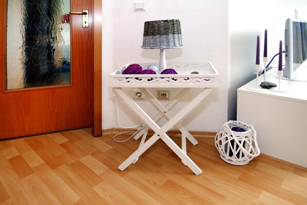 A room with a wooden door, a white ironing board with a basket and purple balls, a white lamp, and a white wall. - Ferienwohnung Kapitänskajute, Dornumersiel (Dornumersiel)