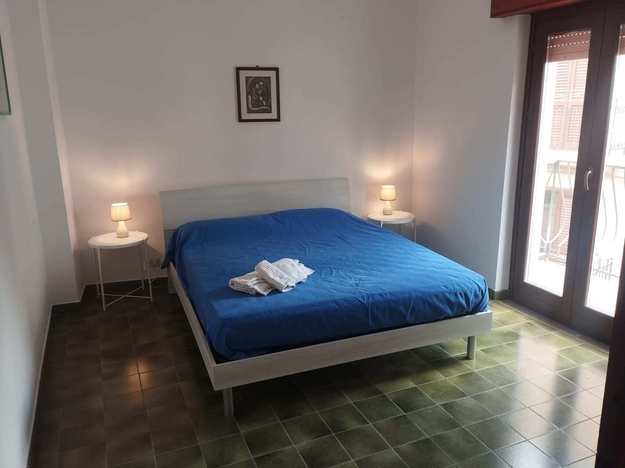 Une chambre à coucher avec un lit, deux tables de chevet avec des lampes, un tableau encadré au mur et une fenêtre donnant sur un paysage.