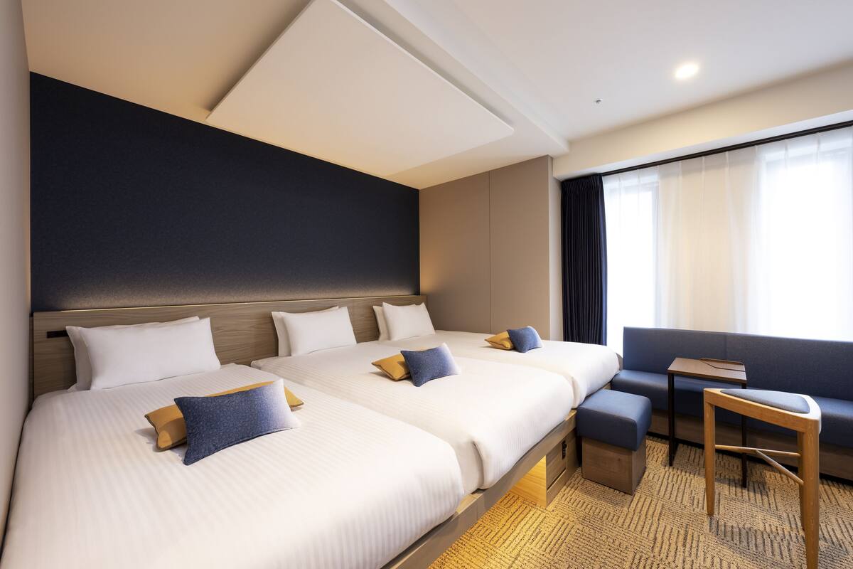 Une chambre d’hôtel avec trois lits, un canapé, une petite table et une grande fenêtre avec des rideaux.